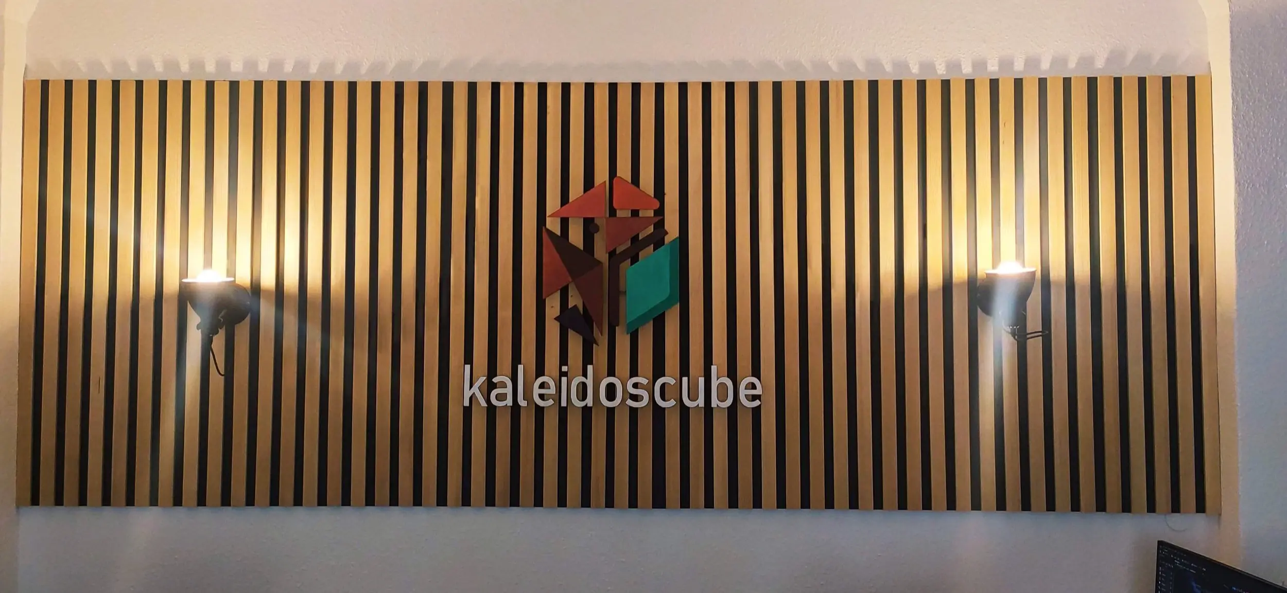 The kaleidoscube logo on a lit wooden slat wall at the studio entrance
