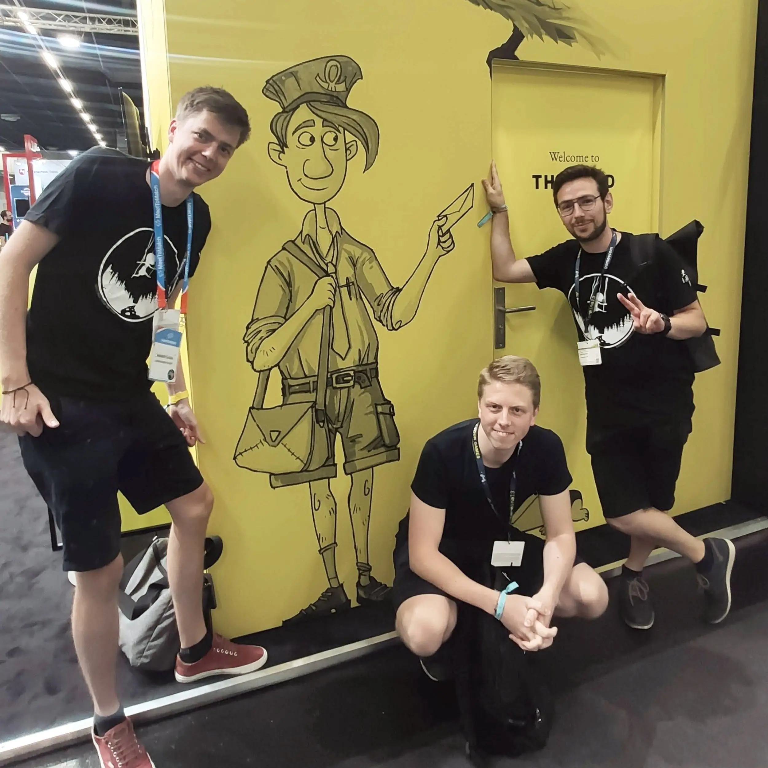 Three team members at a convention beside a wall painted with a character from A Juggler’s Tale
