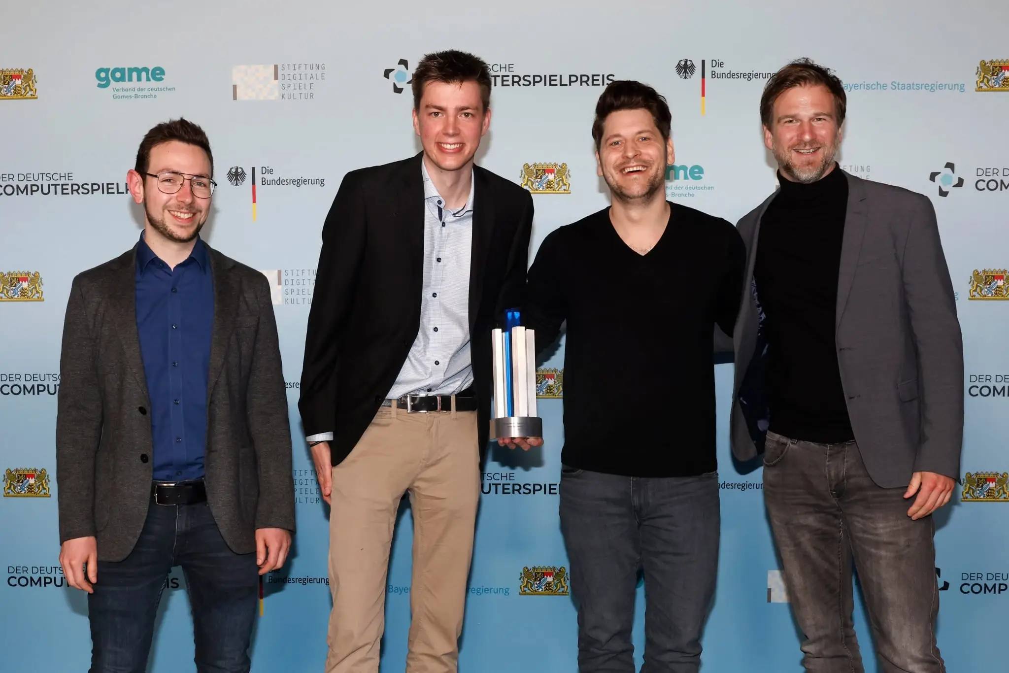 Four team members with the Deutscher Computerspielpreis trophy in front of the press wall